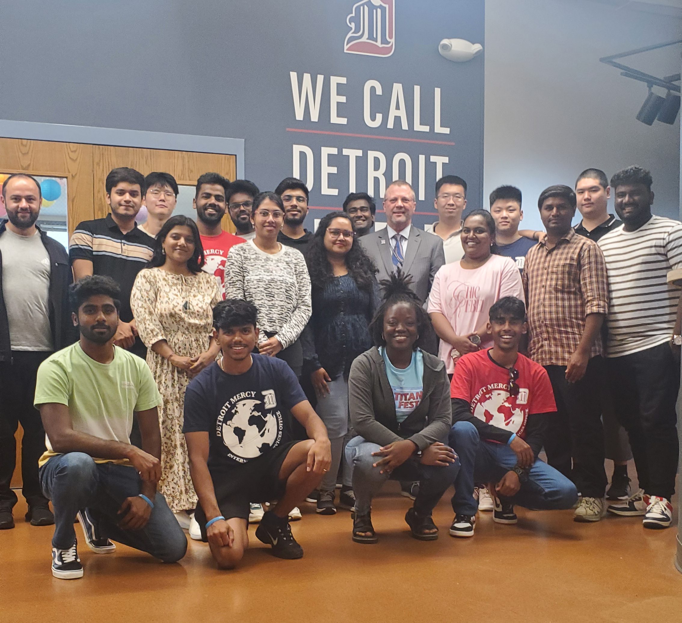 A group of students and President Taylor pose for a photo inside of the Engineering Building, with a large WE CALL DETROIT HOME sign on the wall behind them.