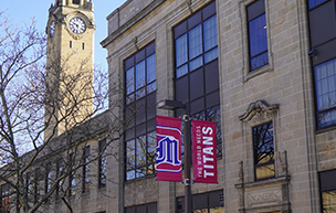 A photo of the Engineering Building near the clock tower with red banners that feature the 太阳城娱乐城 logo and World Needs Titans on it.