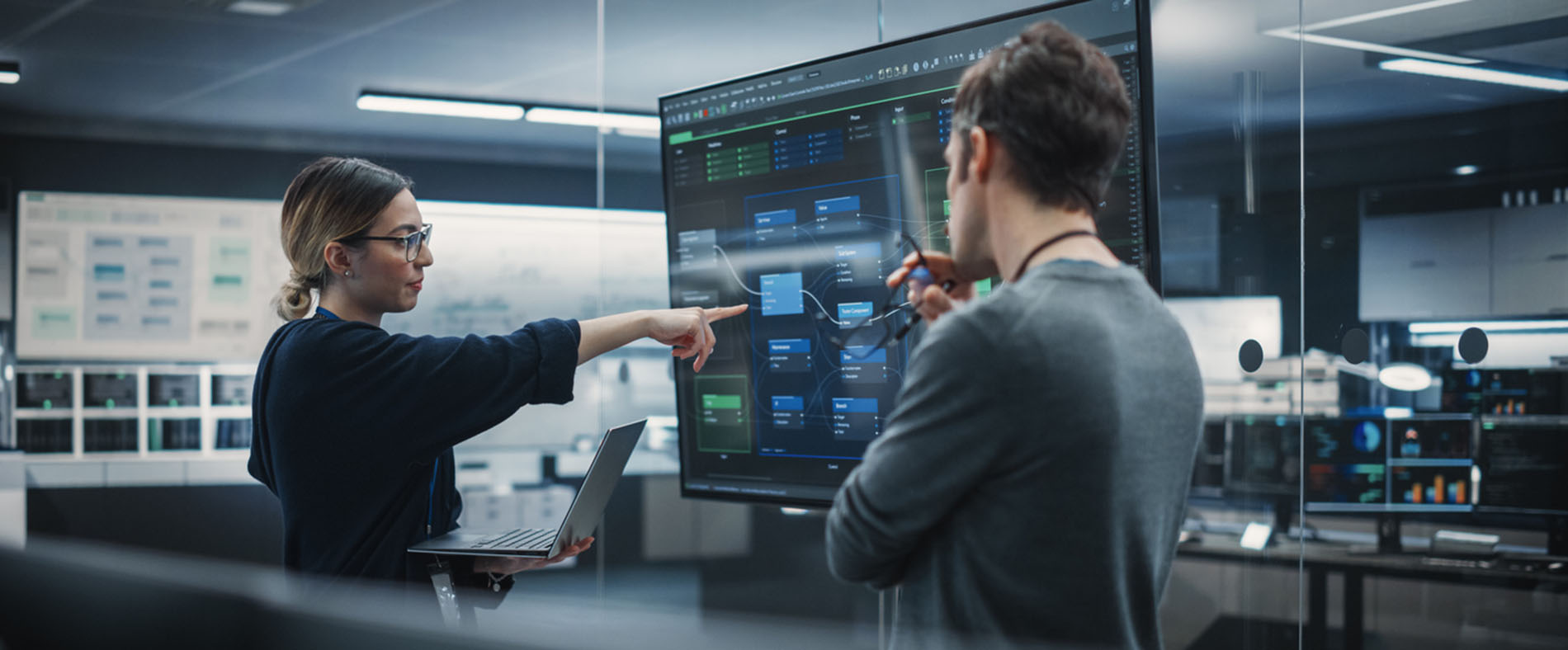 A woman points to a monitor with cybersecurity elements on it as she talks with a man.