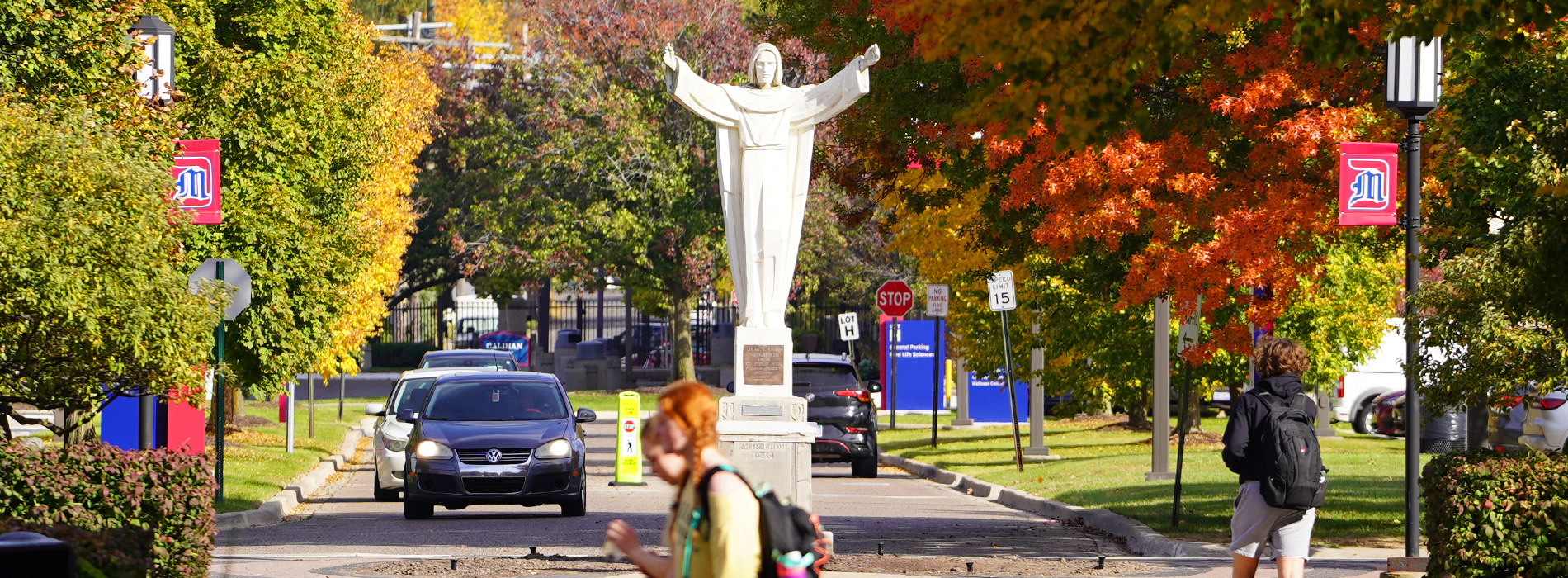 The Jesus statue with students, cars, lamp posts and colorful trees on the McNichols Campus.