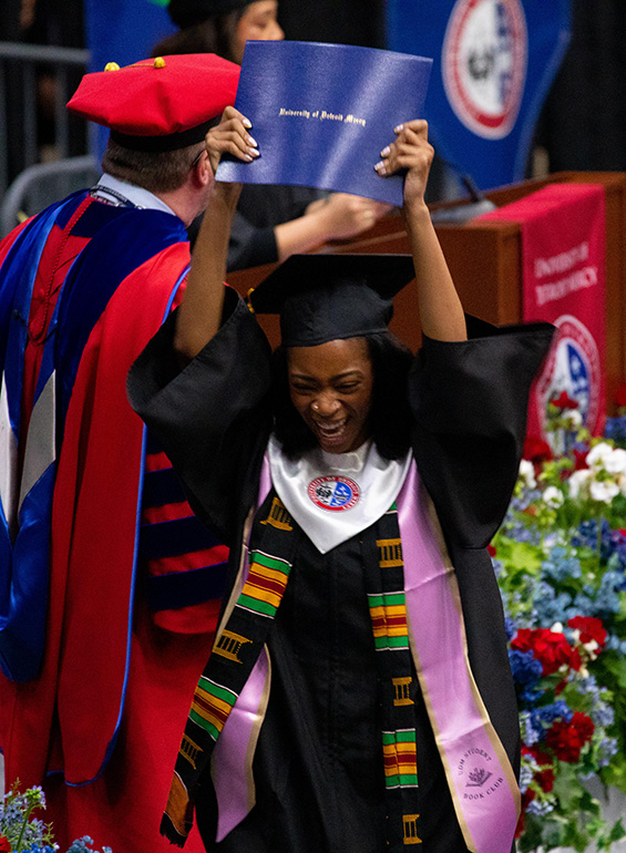 A graduate celebrates by throwing her hands in the air while walking across the Commencement stage.