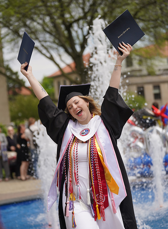 A graduate celebrates with glee by the fountain.