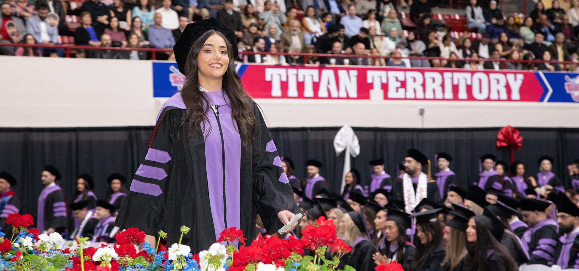 A graduate walks across the stage at Commencement with the Titan Territory sign visible in the background where other graduates are lining up.