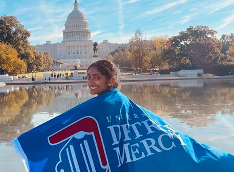 a student standing before the Capitol building with a Detroit Mercy blanket/flag over her shoulders